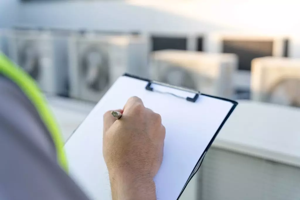 HVAC technician taking notes during routine maintenance check, clipboard in hand with air conditioning units in background